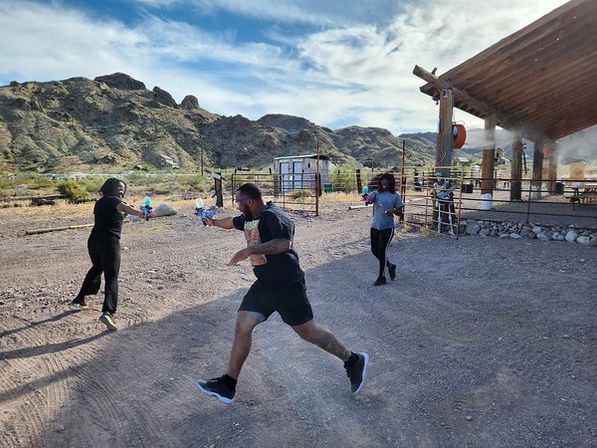 Three people in athletic clothes playfully chase each other with water guns near a wooden pavilion in a sunlit desert mountain landscape