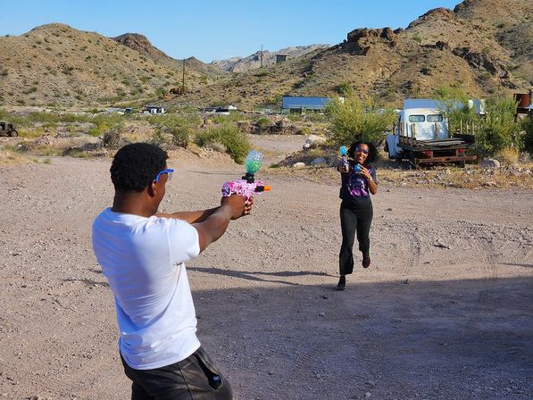 Two people playfully aim colorful toy water guns at each other in a sunny desert gravel lot with rocky hills and an old pickup truck in the background.