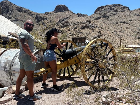 Two visitors examine an antique yellow-wheeled cannon in a sunbaked desert ghost town, with rocky hills, scrub brush and weathered buildings under a clear blue sky.