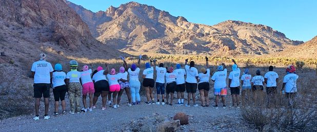 Hiking group in matching white shirts seen from behind, raising hands on a gravel trail in a sunlit desert canyon with rugged rocky mountains and a clear blue sky.