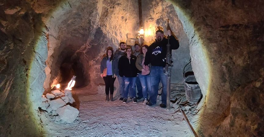 Group of people posing in an illuminated underground mine tunnel with rough rock walls, lanterns, piled stones and vintage mining equipment