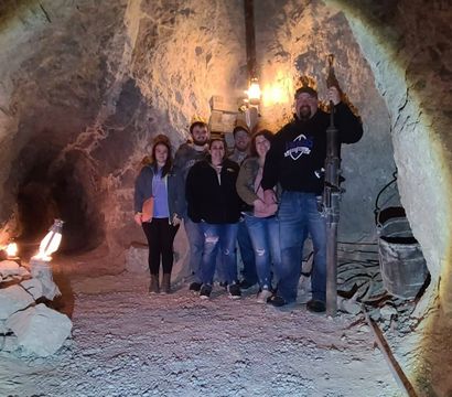 Group of people posing in an illuminated underground mine tunnel with rough rock walls, lanterns, piled stones and vintage mining equipment