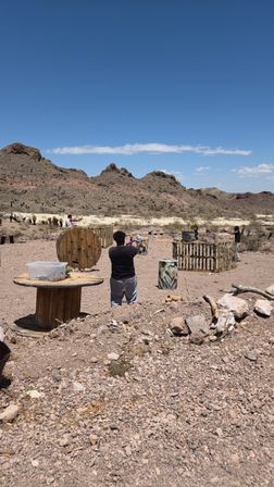 Three people playing a foam-blaster game behind wooden barriers in an arid, rocky desert with craggy hills and a clear blue sky.