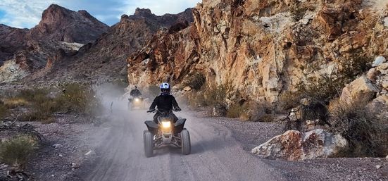 Two helmeted ATV riders on a dusty off-road desert canyon trail, headlights glowing as they kick up dust against rugged rocky cliffs.