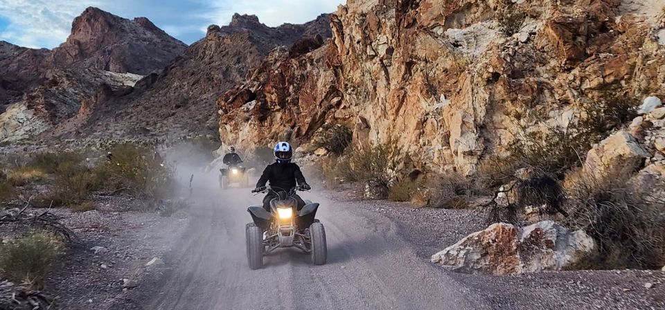 Two helmeted ATV riders on a dusty off-road desert canyon trail, headlights glowing as they kick up dust against rugged rocky cliffs.