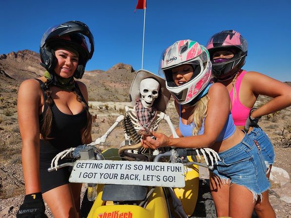 Three helmeted riders in summer outfits pose with a skeleton seated on a yellow ATV in a rocky desert; a sign on the vehicle reads 'Getting dirty is so much fun! I got your back, let's go!'