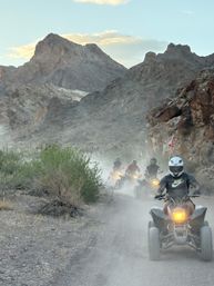 Helmets-on ATV riders kicking up dust on a narrow desert canyon trail with rugged rocky mountains and a pastel evening sky