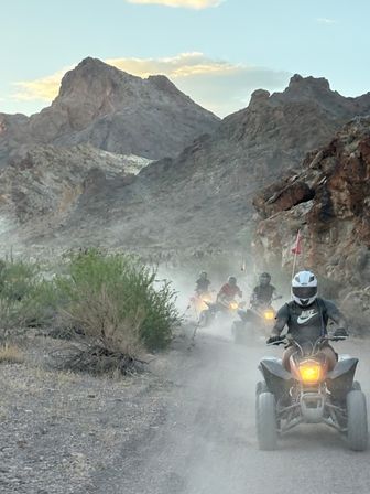Helmets-on ATV riders kicking up dust on a narrow desert canyon trail with rugged rocky mountains and a pastel evening sky