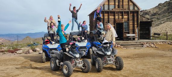 Bachelorette group cheering atop blue off-road ATVs and quads in front of a rustic wooden general store in a desert ghost-town with rocky hills and distant mountains