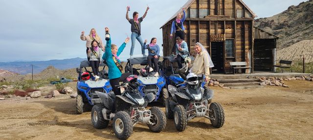 Bachelorette group cheering atop blue off-road ATVs and quads in front of a rustic wooden general store in a desert ghost-town with rocky hills and distant mountains
