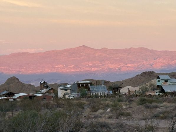 Southwestern desert ghost town with abandoned mining buildings in foreground and pink alpenglow on distant mountains at sunset.