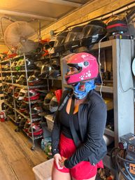 Person wearing a bright pink full-face motorcycle helmet and blue neck gaiter stands inside an indoor helmet storage area with metal racks of assorted helmets, a large industrial fan, wooden floorboards and a workbench with gear.