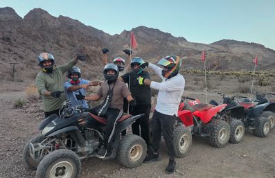 Five helmeted riders posing on and beside red and black ATVs in a rocky desert canyon, off-road quad bikes lined up against a backdrop of rugged mountains and red safety flags.