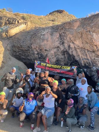 Large group celebrating a birthday at the entrance of a rocky cave on a sunlit hillside, smiling and holding a colorful "Birthday Crew" banner during an outdoor desert hike.