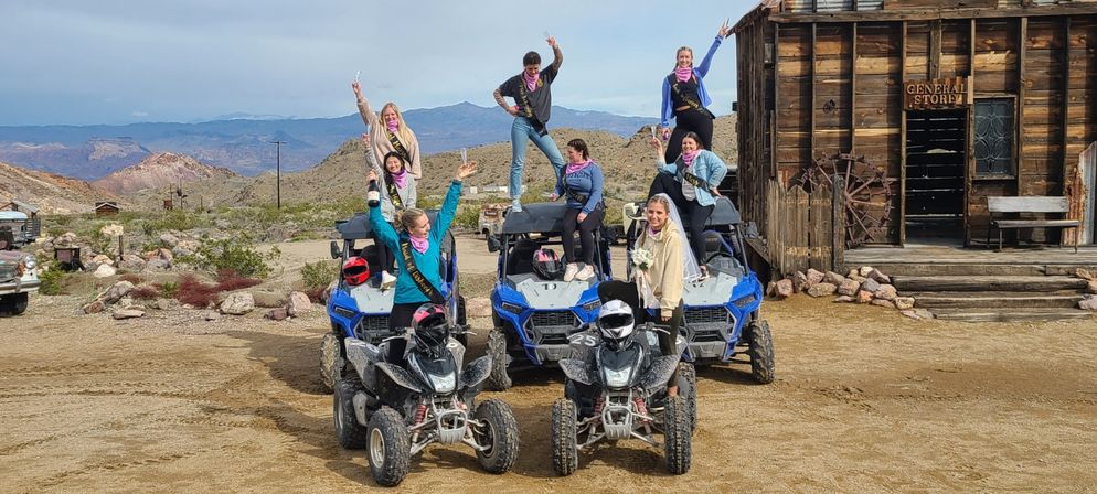 Cheerful bachelorette group in pink bandanas and sashes posing on ATVs and blue UTVs in a desert ghost‑town setting with rugged mountains and a rustic wooden general store.