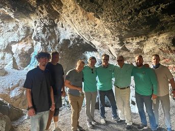 Eight adults in casual hiking clothes pose together inside a sunlit rocky cave entrance during a group outdoor cave tour and adventure.