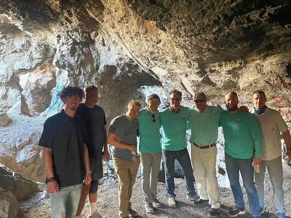Eight adults in casual hiking clothes pose together inside a sunlit rocky cave entrance during a group outdoor cave tour and adventure.