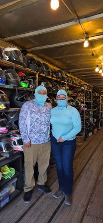 Two people in light-blue balaclavas stand in an aisle of a helmet rental shop, surrounded by rows of motorcycle helmets on wooden shelves and warm hanging bulbs above.
