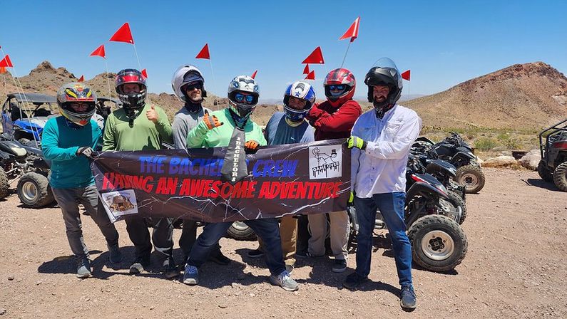 Helmeted ATV riders giving thumbs-up while holding a banner that reads 'Having an Awesome Adventure,' parked with red safety flags against a sunny rocky desert backdrop.