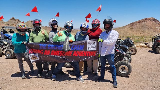 Helmeted ATV riders giving thumbs-up while holding a banner that reads 'Having an Awesome Adventure,' parked with red safety flags against a sunny rocky desert backdrop.