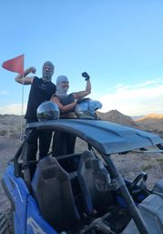 Two people in balaclavas striking a playful victorious pose while standing in a blue off-road UTV, helmets on the roof, on a rocky desert trail with mountains under a clear blue sky.