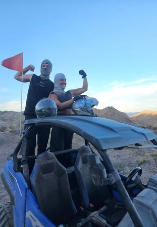 Two people in balaclavas striking a playful victorious pose while standing in a blue off-road UTV, helmets on the roof, on a rocky desert trail with mountains under a clear blue sky.