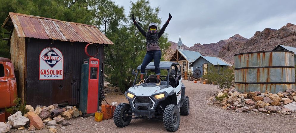 Helmeted rider standing on a white off-road UTV in a desert ghost-town scene with a vintage gas pump, corrugated metal sheds, a small church, water tank, and rocky mountain backdrop.
