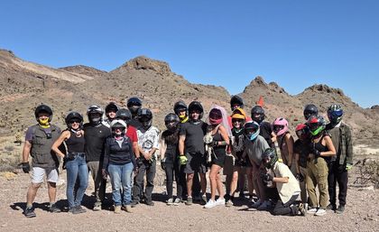 Group of off-road riders in helmets posing with a bride in a pink veil holding a bouquet in a rocky desert mountain landscape under a clear blue sky.