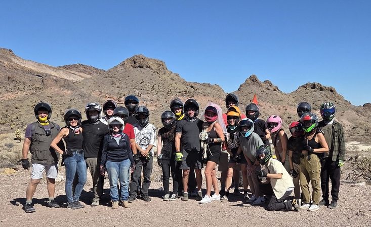 Group of off-road riders in helmets posing with a bride in a pink veil holding a bouquet in a rocky desert mountain landscape under a clear blue sky.