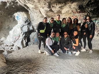 Smiling group of hikers in black activewear and green bandanas posing inside a graffiti-covered rocky cave during an outdoor adventure