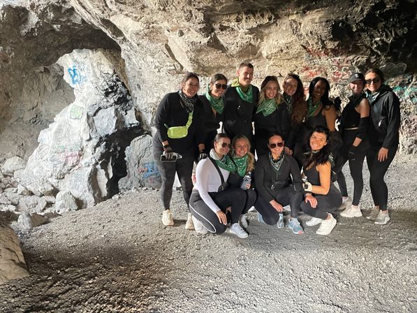 Smiling group of hikers in black activewear and green bandanas posing inside a graffiti-covered rocky cave during an outdoor adventure