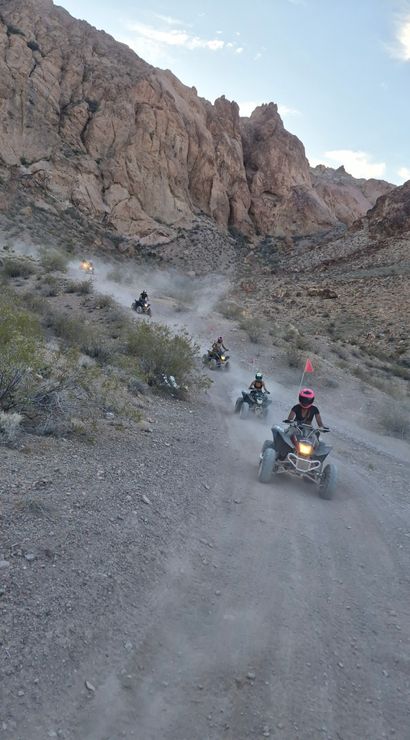 Group of ATV riders on a dusty desert canyon dirt trail, quads kicking up clouds of dust beneath towering rocky cliffs — off‑road adventure in a rugged arid landscape.