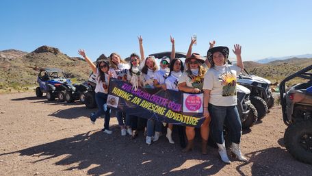 Cheerful bachelorette crew cheering in front of UTVs and ATVs on a rocky desert trail, holding a colorful banner with arid hills and clear blue sky behind them.