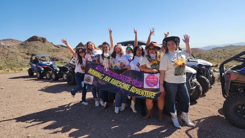 Cheerful bachelorette crew cheering in front of UTVs and ATVs on a rocky desert trail, holding a colorful banner with arid hills and clear blue sky behind them.