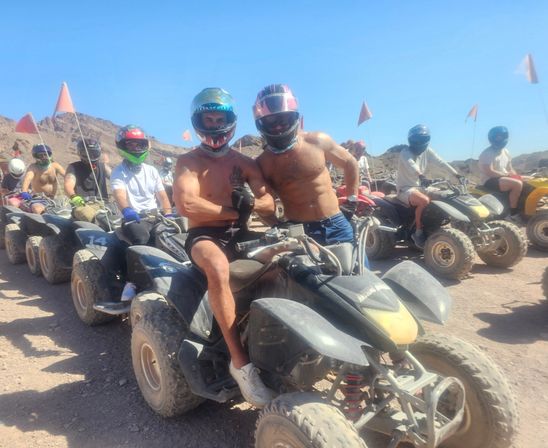 Line of helmeted riders on ATVs in a sunny rocky desert, two riders posing on front quads.