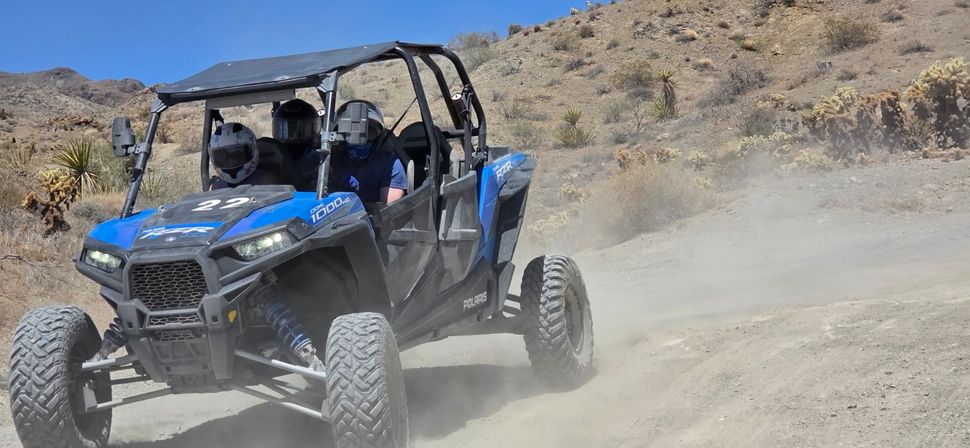Blue side-by-side UTV tearing along a dusty desert trail with helmeted riders, rocky hills and scattered cacti in the background.