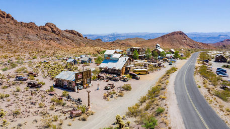Aerial view of a desert ghost town with weathered wooden buildings, rusted vintage cars and a yellow school bus beside a highway winding through rocky hills.