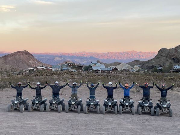 Row of ATV riders in helmets cheering with arms raised on quad bikes in a desert foreground, pastel sunset lighting distant mountains and a rustic ghost-town of old mining buildings.