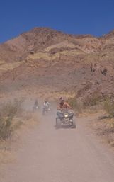 Three ATV riders kicking up dust on a sunny desert trail beneath layered rocky mountains and a clear blue sky.