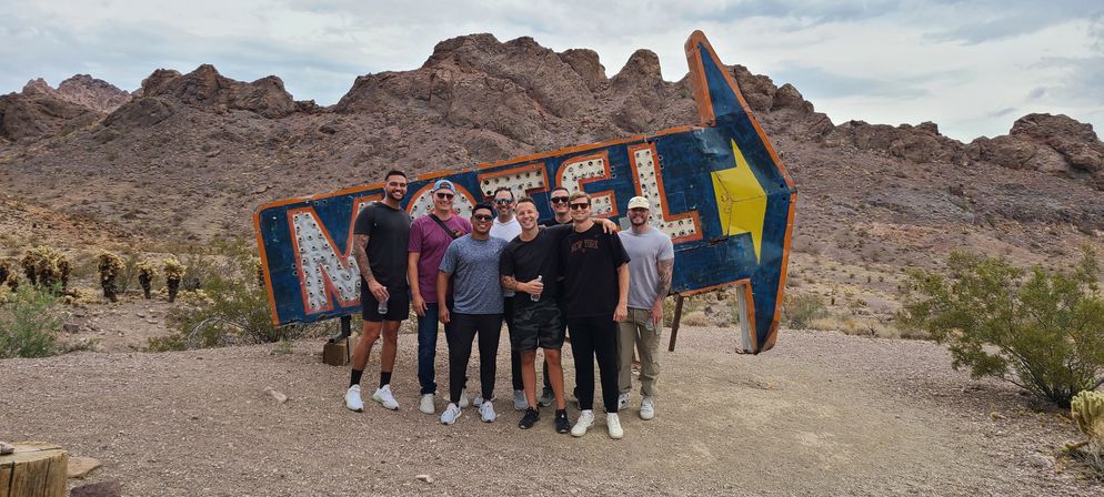 Eight people smiling and posing in front of a weathered vintage motel arrow sign with a yellow star, set against rugged rocky hills in a dry desert landscape under a cloudy sky.