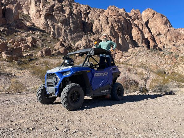 Blue off-road UTV parked on a desert trail with a person standing through the roll cage, surrounded by red rock cliffs and a clear blue sky.