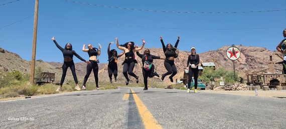Group of friends in black activewear jumping across a sunlit desert highway with red rocky hills, rustic buildings and a retro gas-station sign in the background under a clear blue sky.