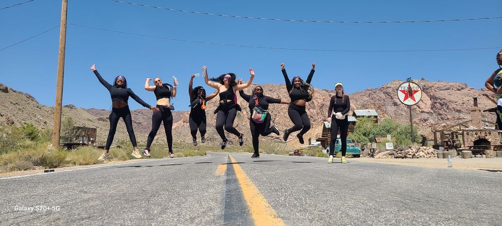 Group of friends in black activewear jumping across a sunlit desert highway with red rocky hills, rustic buildings and a retro gas-station sign in the background under a clear blue sky.