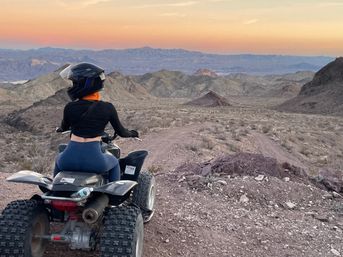 Person in a helmet and orange neck gaiter sitting on an ATV, overlooking rocky desert mountains and a winding off‑road trail at sunset with a pastel sky.