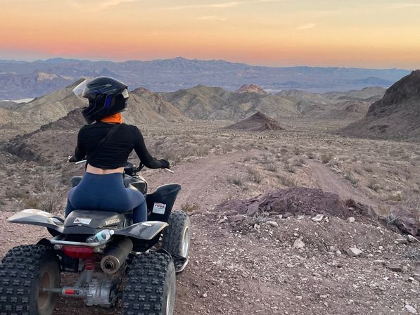 Person in a helmet and orange neck gaiter sitting on an ATV, overlooking rocky desert mountains and a winding off‑road trail at sunset with a pastel sky.