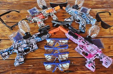 Four colorful gel‑blaster toy guns with clear bead reservoirs and orange muzzles, assorted safety goggles and sunglasses arranged on a rustic wooden table — playful backyard party gear.