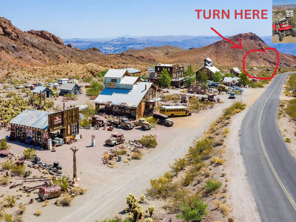 Aerial view of a quirky desert ghost town roadside attraction with weathered wooden buildings, vintage cars and a yellow school bus beside a paved highway and arid red-rock mountains, with a red handwritten “TURN HERE” arrow pointing to a dirt turnoff.