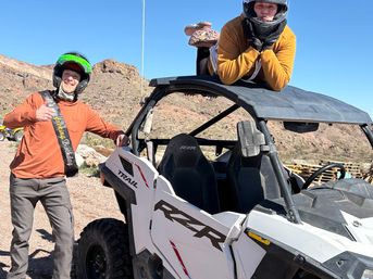 Two helmeted riders posing with a white side-by-side off-road UTV on a sunny desert trail — one giving a thumbs-up wearing a birthday sash and the other lounging on the vehicle roof with rocky canyon hills and a clear blue sky behind them.