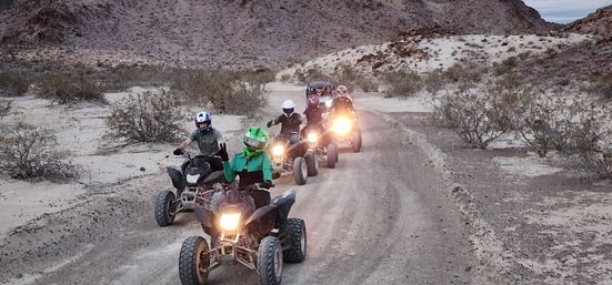 Five helmeted riders on ATVs (quads) riding a dirt desert trail with headlights on, surrounded by scrub and rocky hills.