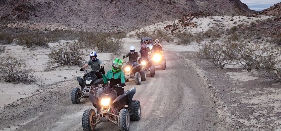 Five helmeted riders on ATVs (quads) riding a dirt desert trail with headlights on, surrounded by scrub and rocky hills.